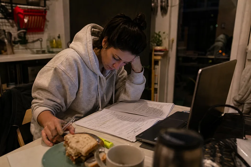 Woman in a hoodie reading a printed project quote at her kitchen table late in the evening, laptop open beside her