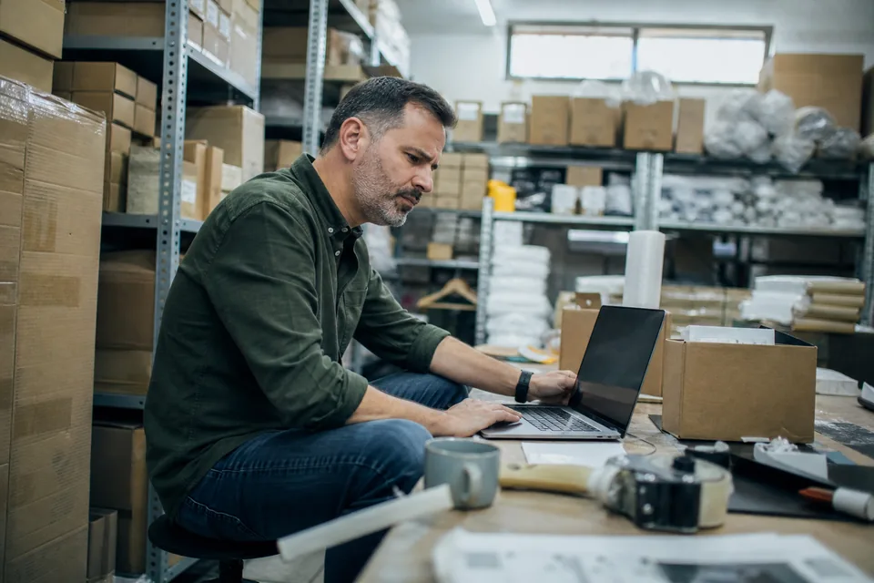 Online retailer reviewing a product page on a laptop in a cluttered stockroom between shelves of boxed inventory