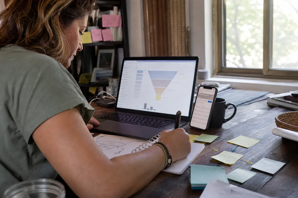 Woman reviewing a sales funnel on her laptop and phone with a hand-drawn funnel sketch and sticky notes on the desk