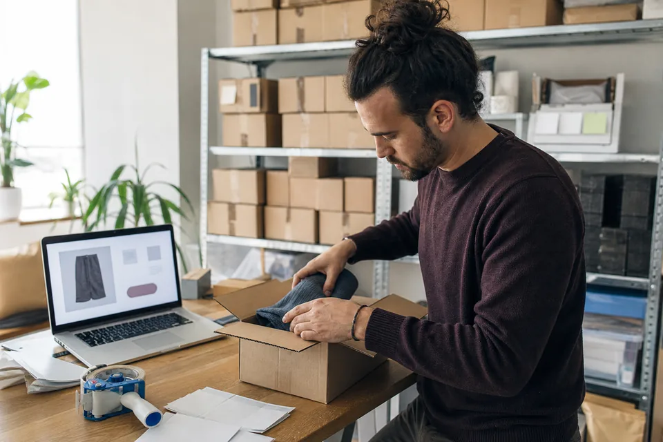 Online store owner packing an order beside a laptop showing a product page and checkout